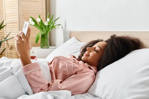 Una mujer afroamericana rizada en pijama yace en la cama, sosteniendo un teléfono celular — Stock Photo