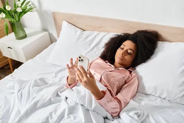 Curly African American woman in pajamas relaxes on bed, holding cell phone in morning light. — Stock Photo