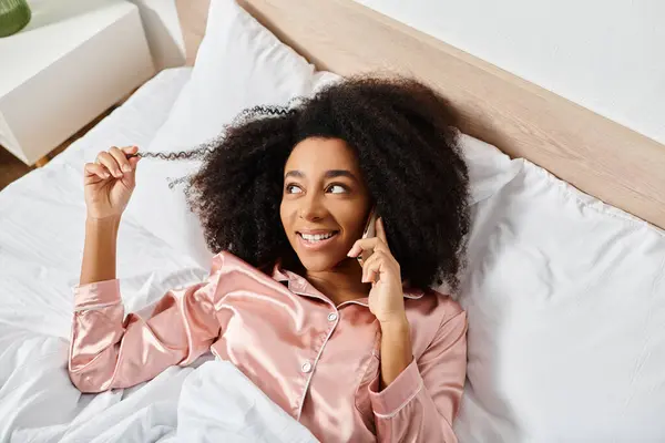 Curly African American woman in pajamas, laying in bed, talking on a cell phone in a cozy bedroom setting. — Stock Photo