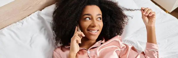 Curly African American woman in pajamas lying in bed, having a conversation on her cell phone in the morning. — Stock Photo