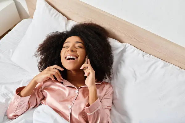 Una mujer afroamericana rizada en pijama se acuesta en la cama, charlando en su teléfono celular a la luz de la mañana. - foto de stock