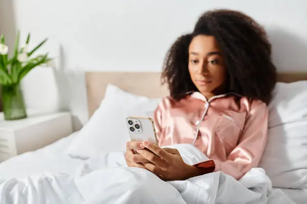 Curly African American woman in pajamas laying in bed, engrossed with cell phone in early morning light. — Stock Photo