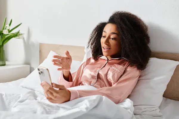 A curly African American woman in pajamas sits on a bed, engrossed in a tablet in a cozy bedroom setting. — Stock Photo