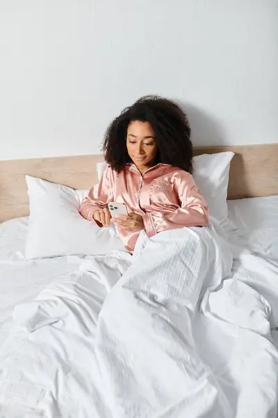 Riccio donna afroamericana in pigiama sdraiato a letto, assorbito nel suo schermo del telefono cellulare al mattino. — Foto stock