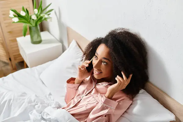 Mujer afroamericana rizada en pijama hablando por teléfono celular mientras está acostada en la cama en la mañana. - foto de stock
