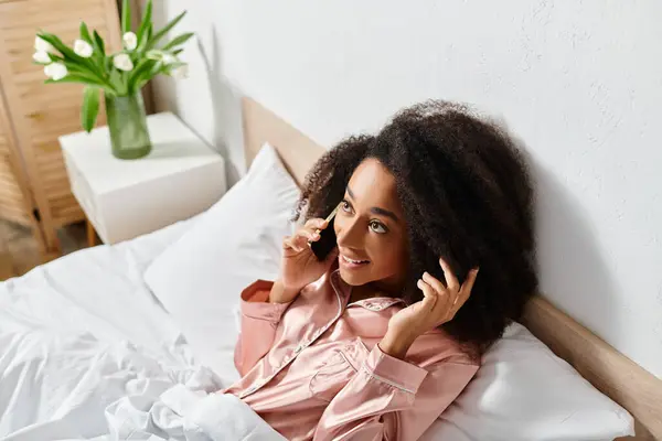 Curly African American woman in pajamas talking on cell phone while laying on bed in bedroom in the morning. — Stock Photo