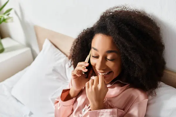 Curly African American woman in pajamas talking animatedly on cell phone while lounging in bed in the morning. — Stock Photo