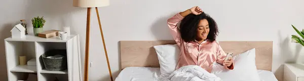 Una mujer afroamericana rizada se relaja en la parte superior de una cama en su dormitorio, vistiendo una camisa rosa durante la tranquila mañana. - foto de stock