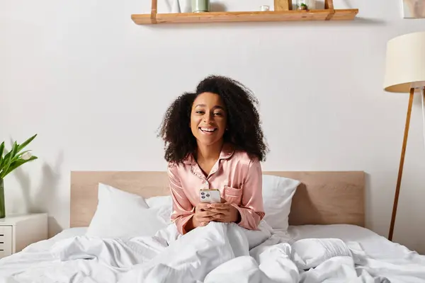 Donna afroamericana riccia in pigiama, seduta sul letto, assorta nel cellulare. Filtri di luce del mattino attraverso la finestra. — Foto stock