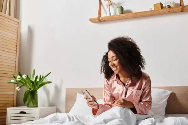 Curly African American woman in pajamas sits on bed, deep in thought, focusing on her cell phone screen in the morning. — Stock Photo