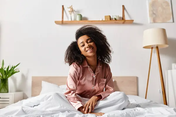 A curly African American woman in pajamas sits atop a bed, smiling brightly in the morning light. - foto de stock