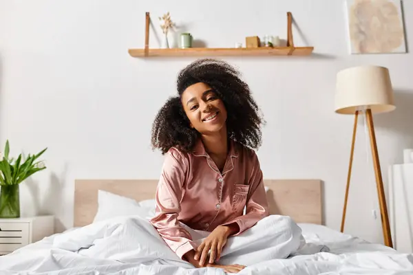 A curly African American woman in pajamas sits atop a bed, smiling brightly in the morning light. — Stock Photo