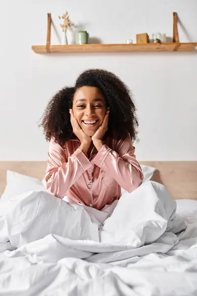 Curly African American woman in pajamas relaxing on a white sheet-covered bed in the morning. — Stock Photo