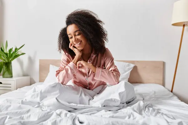 A curly African American woman in pajamas sits calmly on a bed with white sheets in a serene bedroom in the morning. — Stock Photo