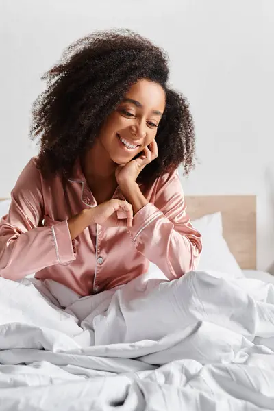 Uma mulher afro-americana encaracolada vestida de pijama fica tranquilamente em uma cama com lençóis brancos em um quarto aconchegante durante o horário da manhã.. — Fotografia de Stock