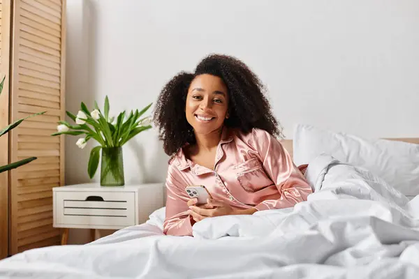Curly African American woman in pajamas sitting on bed, holding cell phone in the morning. — Stock Photo