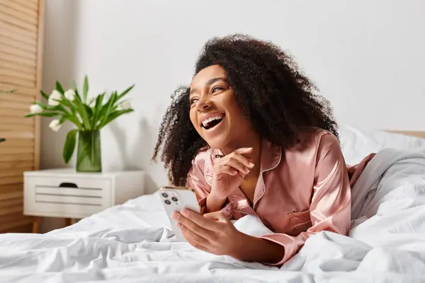 A curly African American woman, dressed in pajamas, peacefully laying on a bed while holding a cell phone. — Stock Photo