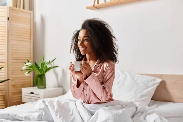 A curly African American woman in pajamas sitting on a bed, enjoying a cup of coffee in the morning sunlight. — Stock Photo