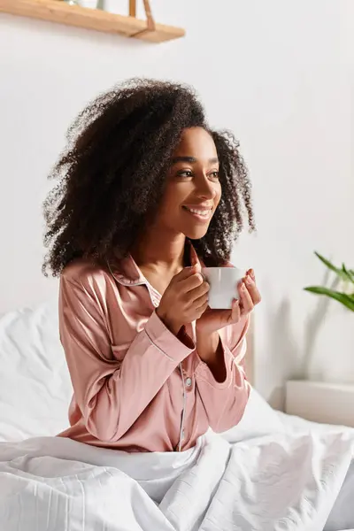 Riccio donna afroamericana in pigiama si siede su un letto, tenendo una tazza di caffè in una camera da letto accogliente durante il mattino. — Foto stock