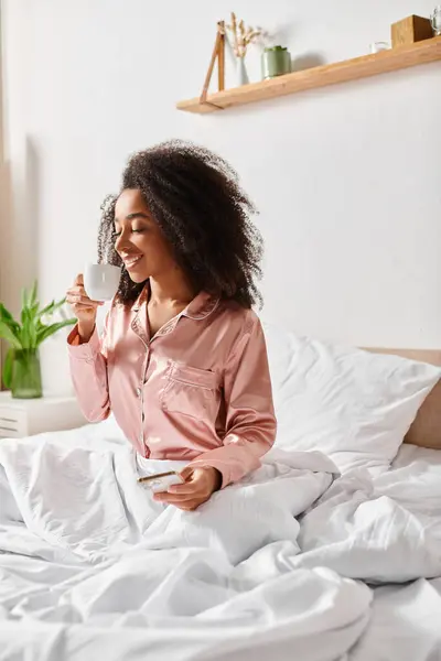 Una mujer afroamericana con el pelo rizado se sienta en una cama, sosteniendo una taza en un ambiente acogedor dormitorio durante la mañana. - foto de stock