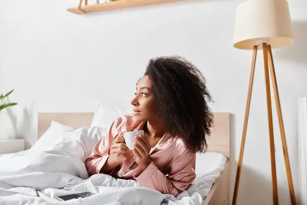 Una donna afroamericana riccia in pigiama si rilassa a letto con una tazza di caffè durante la mattina. — Foto stock