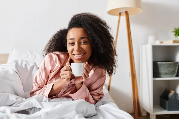 Femme afro-américaine frisée en pyjama, nichée au lit, savourant sa matinée avec une tasse de café. — Photo de stock