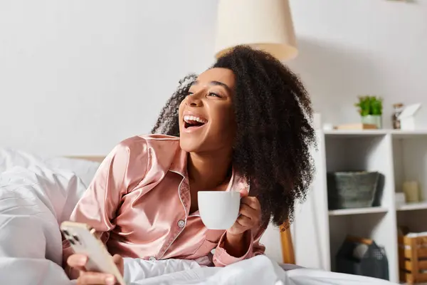 A curly African American woman in pajamas relaxing in bed with a cup of coffee in a cozy bedroom during morning time. — Stock Photo