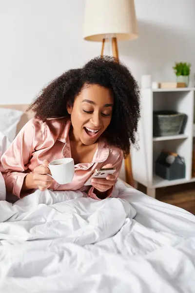 Riccio donna afroamericana in pigiama rilassante a letto, assaporando una tazza di caffè durante le ore del mattino. — Foto stock