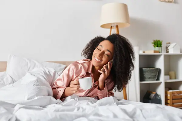 Una mujer afroamericana con el pelo rizado en pijama, saboreando una taza de café en un acogedor dormitorio durante la mañana. - foto de stock