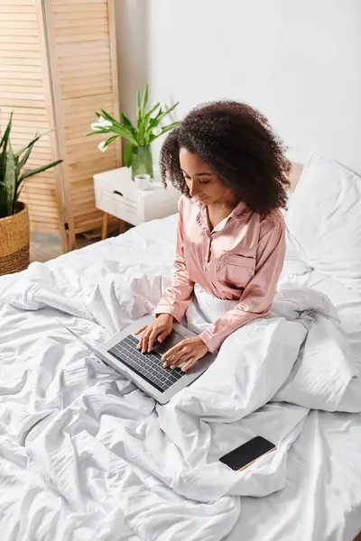 Curly African American woman in pajamas sitting on bed, focused on laptop screen, in cozy bedroom ambiance. — Stock Photo