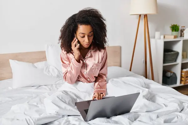Curly African American woman in pajamas sits on bed, focused on using laptop in cozy bedroom during morning time. — Stock Photo