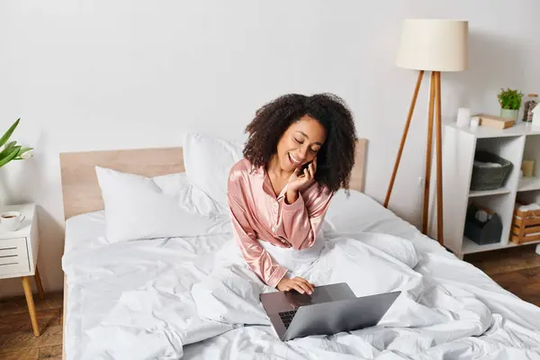 Curly African American woman in pajamas sits on bed, absorbed in laptop screen, in cozy bedroom setting during morning time. — Stock Photo