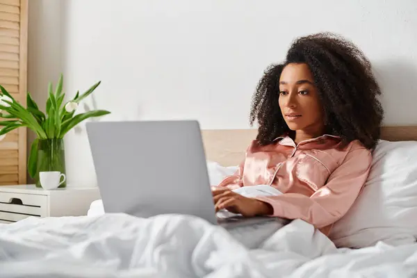 Una donna afroamericana riccia in pigiama si siede su un letto, assorbita nel lavorare su un computer portatile al mattino. — Foto stock