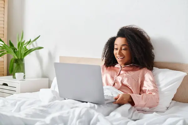 African American woman in pajamas sitting on bed, using laptop in bedroom in the morning. — Stock Photo
