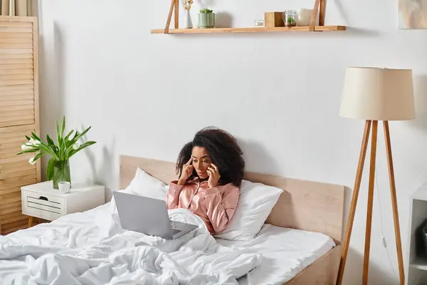 Una tranquilla scena mattutina con una donna afroamericana riccia in pigiama seduta su un letto, concentrata sull'utilizzo del suo computer portatile. — Foto stock