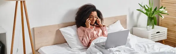 Una donna afroamericana riccia in pigiama si siede su un letto, assorbita dall'uso di un computer portatile al mattino. — Foto stock