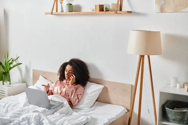 Curly afro-americano mulher de pijama sentado na cama, falando no telefone celular no quarto pela manhã. — Fotografia de Stock