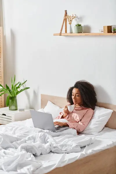 A curly African American woman in pajamas sits on a bed in a bedroom, focused on her laptop screen in the morning light. — Stock Photo