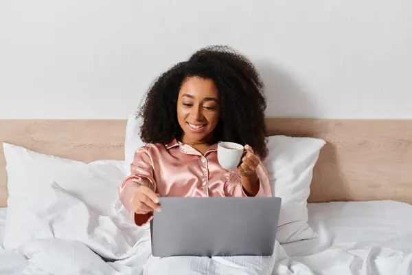 Femme afro-américaine bouclée en pyjama assis sur le lit, tenant une tasse de café, regardant l'écran d'ordinateur portable dans la chambre. — Photo de stock
