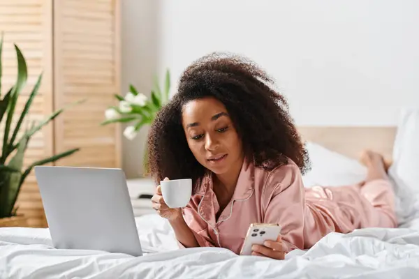 Curly African American woman in pajamas enjoying a leisurely morning with a laptop and a cup of coffee on her bed. — Stock Photo