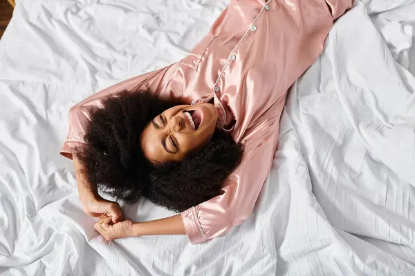 Curly mulher afro-americana vestindo pijama, pacificamente deitado em cima de uma cama branca em seu quarto durante o tempo da manhã. — Fotografia de Stock