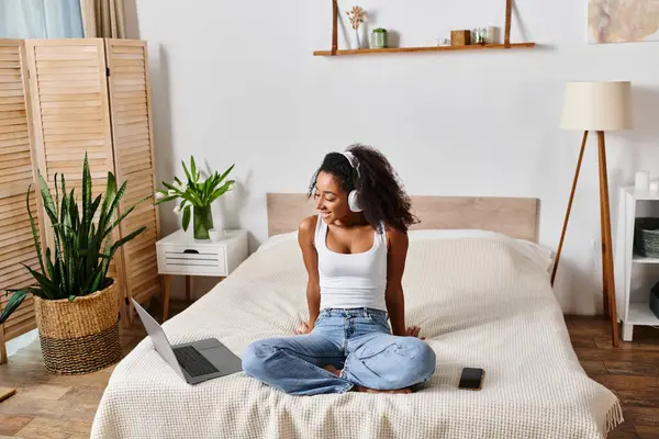 A curly African American woman in a tank top sits on a bed, engrossed in her laptop. — Stock Photo