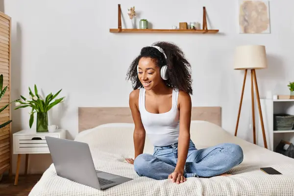 A curly African American woman in a tank top sits on a bed, deeply focused on her laptop in a modern bedroom. — Stock Photo