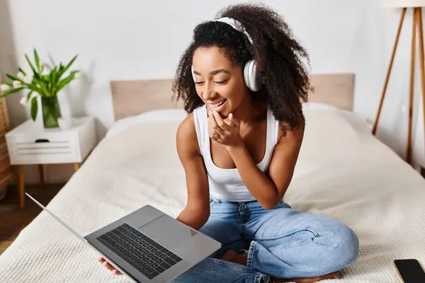 Curly African American woman in tank top sitting on bed, engrossed in laptop screen, in modern bedroom. — Stock Photo