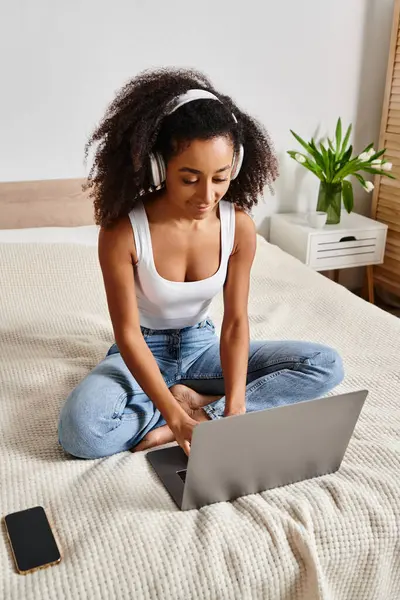 Curly African American woman sitting on bed, typing on laptop in modern bedroom setting. — Stock Photo