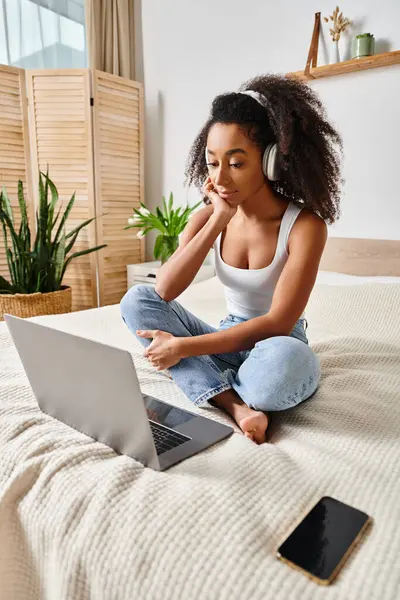 A curly African American woman sits on a bed, focused on her laptop screen in a modern bedroom. — Stock Photo
