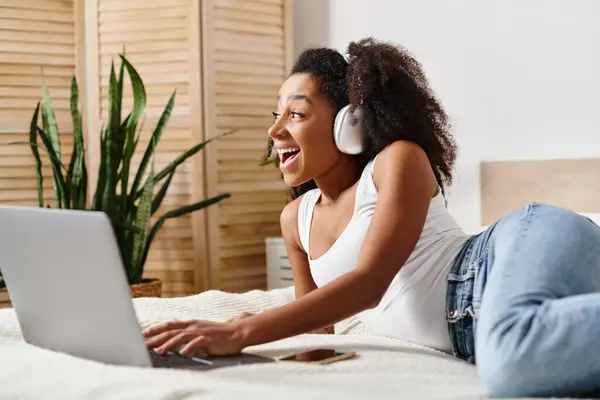 A curly African American woman in a tank top relaxes on a bed, using a laptop computer in a modern bedroom. — Stock Photo