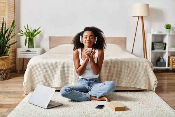 Una mujer afroamericana rizada en una camiseta sin mangas se sienta serenamente en el suelo frente a una cama moderna. - foto de stock