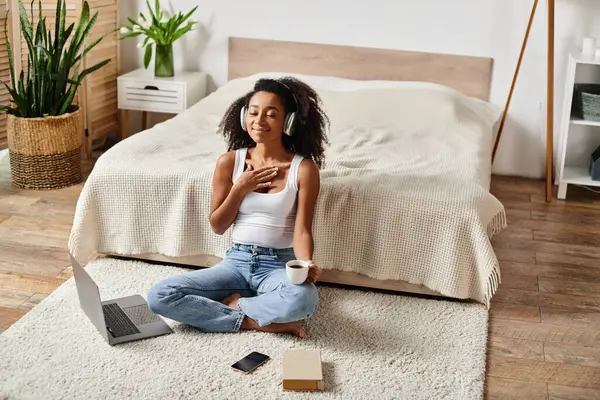 Mujer afroamericana rizada sentada en el suelo en interiores, centrada en el uso de una computadora portátil en un entorno de dormitorio moderno. — Stock Photo