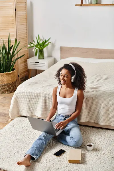 Una donna afroamericana con i capelli ricci si siede sul pavimento in una camera da letto moderna, utilizzando un computer portatile. — Foto stock